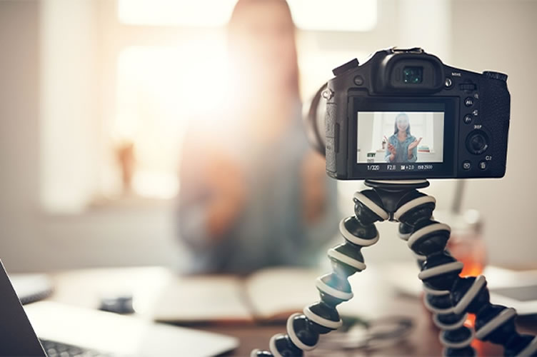No primeiro plano da imagem uma câmera profissional, ao fundo e embraçado uma corretora de imóveis gravando vídeos para anúncios de imóveis