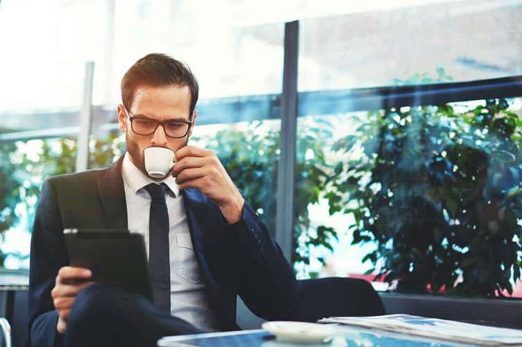Corretor de imóveis de terno tomando um café, com óculos e um tablet nas mãos observando suas redes sociais. 
