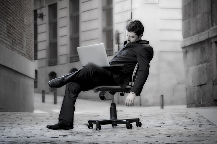 Corretor de imóveis com terno e notebook no colo. Ele está sentado em uma cadeira no meio do nada e exausto por não conseguir clientes para o seu negócio.