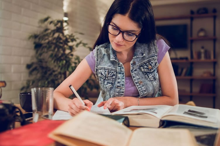 Mulher de cabelo escuro, usando óculos estudando para a faculdade de negócios imobiliários. Diversos livros sob a mesa.