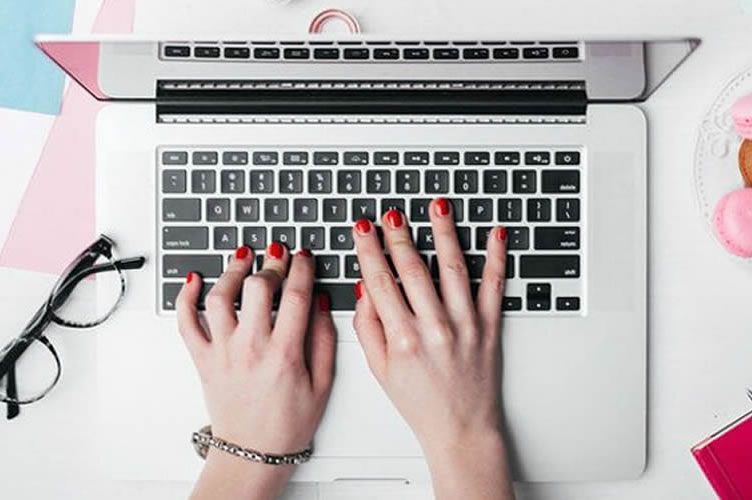 Mostra as mãos de uma corretora de imóveis no teclado de um notebook para começar a criar conteúdo para o blog imobiliário. Ao lado, óculos e no pulso uma pulseira.